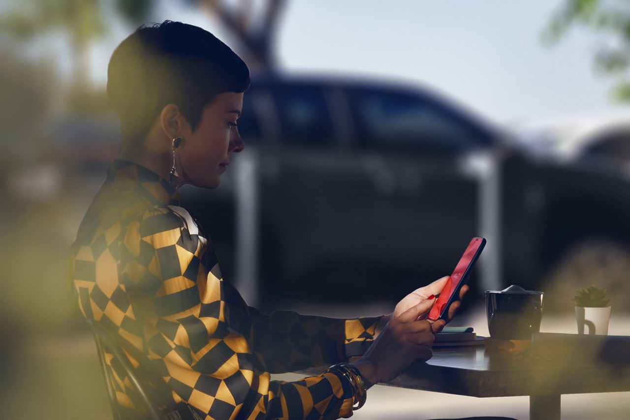 Lady sat at a restaurant table using the TOYOTA MyT app on her phone, her Toyota Yaris Cross is parked in the background.